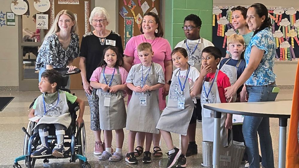 A group photo in a school hallway featuring seven students wearing "Wild Mustang Coffee Co." aprons and four smiling adults. One student in the front is using a wheelchair, and the group is gathered together for a team portrait during their coffee shop program.