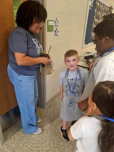 An elementary student wearing a grey "Wild Mustang Coffee Co." apron smiles while interacting with a staff member in a hallway.