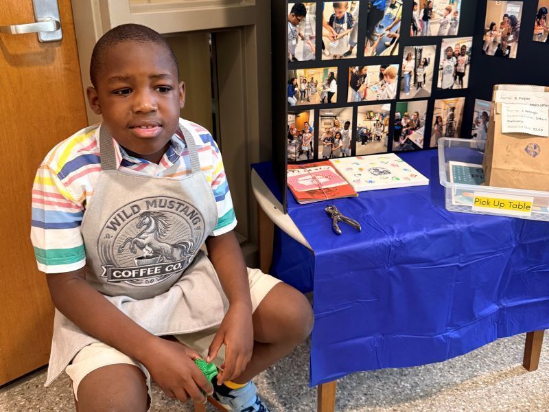 A student in a "Wild Mustang Coffee Co." apron sitting next to an order pick-up table and photo display.